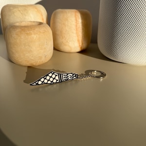 Könnte beinhalten: Ein Schlüsselanhänger mit schwarz-weißem geometrischem Design und einem silbernen Schlüsselring. Der Schlüsselanhänger befindet sich vor drei beige Stein-Kerzenhaltern und einem weißen Lautsprecher.