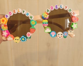 May include: Pink round sunglasses with the name "Adelynn" spelled out in colorful beads around the frames. The sunglasses are decorated with colorful cartoon charms, including flowers, smiley faces, and a rainbow.