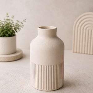Puede incluir: Un jarrón de cerámica color crema con cuerpo cilíndrico y cuello estrecho. La parte inferior del jarrón presenta detalles acanalados verticales. Una pequeña planta en maceta y un arco decorativo están en el fondo. El jarrón es un adorno decorativo para el hogar.