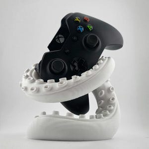 Pode incluir: Um comando de videojogos preto que repousa dentro de um suporte branco em forma de tentáculo de polvo enrolado. O comando apresenta um corpo preto com um logótipo circular Xbox, dois joysticks pretos, um direcional preto e quatro botões de ação coloridos: Y, X, B e A.