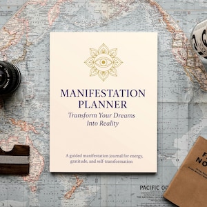 Könnte beinhalten: Ein cremefarbenes "Manifestation Planner" Journal mit goldenen Akzenten und dem Text "Transform Your Dreams Into Reality". Ein Kameraobjektiv, eine Holzgeldbörse und ein "Field Notes" Notizbuch liegen auf einer Vintage-Karte.