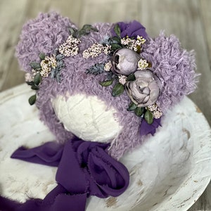 May include: A lavender fuzzy baby hat with a flower crown and a purple satin ribbon. The hat is on a white wooden bowl.