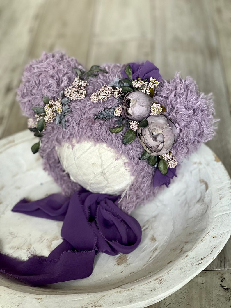 May include: A lavender fuzzy baby hat with a flower crown and a purple satin ribbon. The hat is on a white wooden bowl.