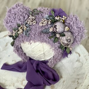 May include: A lavender fuzzy hat with a flower crown and a purple ribbon. The hat is on a white wooden surface.