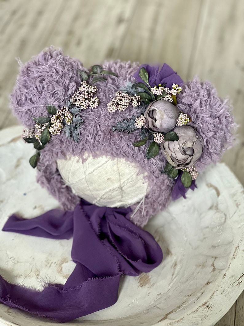 May include: A lavender fuzzy hat with a flower crown and a purple ribbon. The hat is on a white wooden surface.
