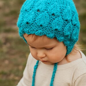 May include: A turquoise knitted baby bonnet with a scalloped edge and leaf-like patterns. The bonnet has long, braided ties. The baby is wearing a cream-colored long-sleeved shirt.