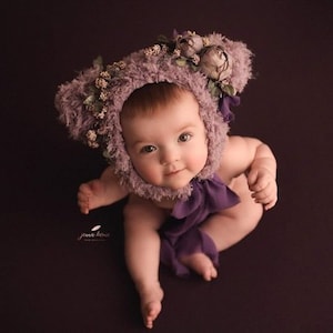 May include: A baby wearing a purple fuzzy hat with flower embellishments. The hat has bear ears. The baby is sitting on a purple background.