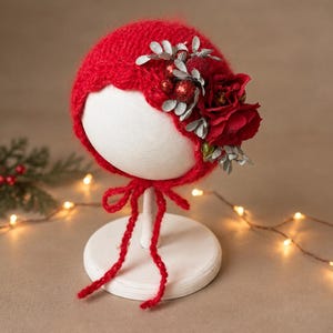 May include: A bright red knitted bonnet featuring a scalloped edge and chin ties. The side is adorned with a cluster of silver-toned foliage, small red glittery spheres, and a large, deep red fabric flower attached to the textured yarn surface.