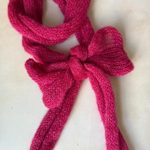 Op de afbeelding: Een levendige fuchsia roze sjaal gemaakt van fijn, semi-transparant gebreid garen. De sjaal is in het midden geknoopt tot een opvallende, decoratieve strik, met twee lange, losse uiteinden die naar beneden hangen en een lus naar boven.