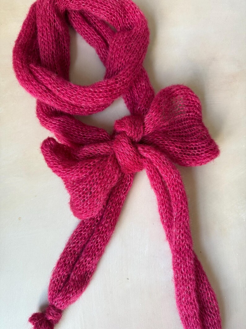 Op de afbeelding: Een levendige fuchsia roze sjaal gemaakt van fijn, semi-transparant gebreid garen. De sjaal is in het midden geknoopt tot een opvallende, decoratieve strik, met twee lange, losse uiteinden die naar beneden hangen en een lus naar boven.