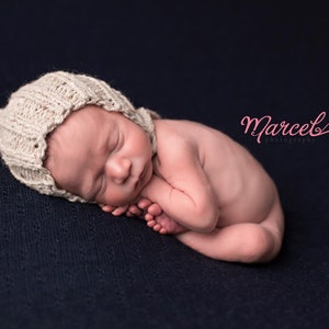 May include: A newborn baby wearing a beige knitted bonnet, curled up on a dark blue textured surface. The baby is sleeping peacefully, with tiny feet visible. The image includes the text "Marcel's Photography".
