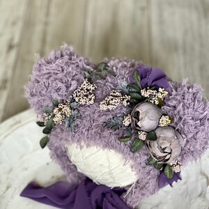 May include: A purple fuzzy hat with a flower crown and a matching purple ribbon. The hat is made of soft, fluffy material and is perfect for newborn photography.