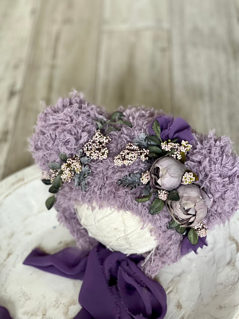 May include: A purple fuzzy hat with a flower crown and a matching purple ribbon. The hat is made of soft, fluffy material and is perfect for newborn photography.