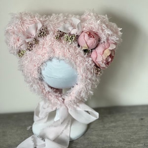 May include: A pink fuzzy hat with bear ears and pink flowers. The hat is on a white stand with a pink ribbon.
