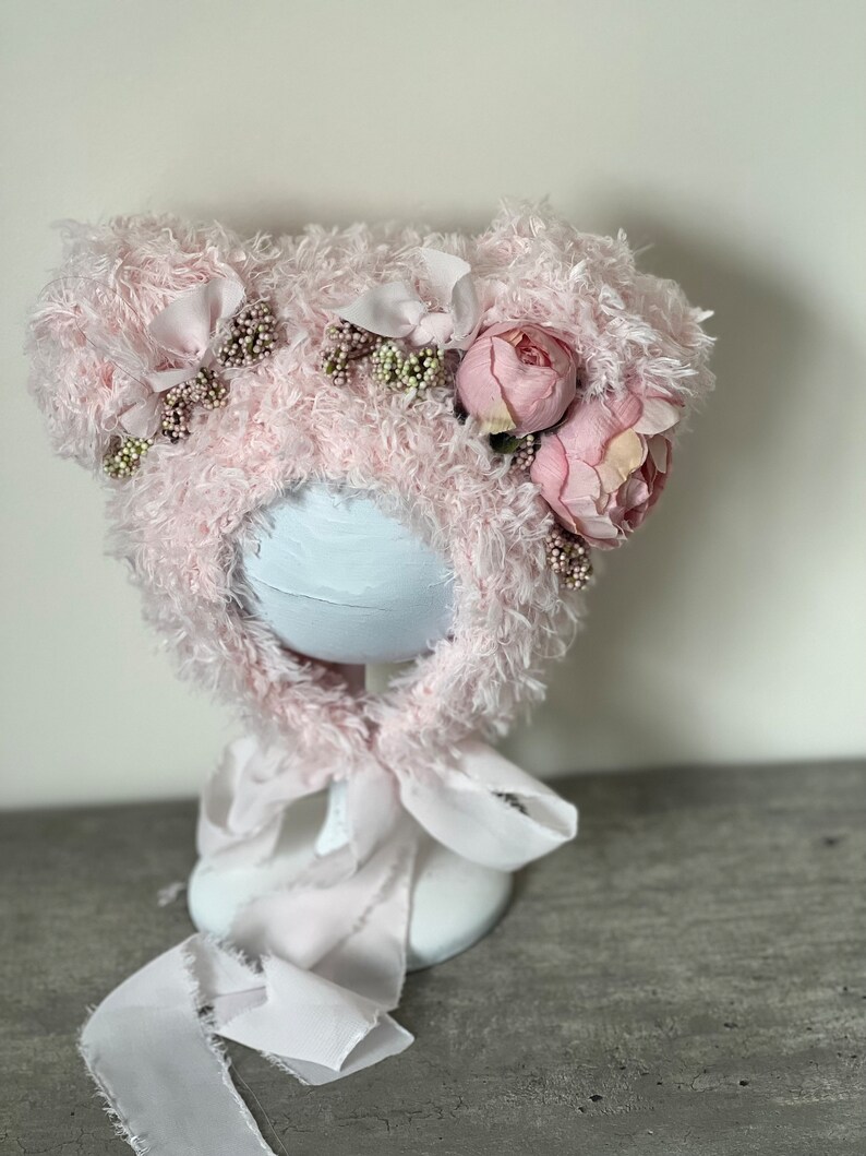 May include: A pink fuzzy hat with bear ears and pink flowers. The hat is on a white stand with a pink ribbon.