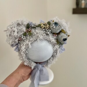 May include: A gray fuzzy baby hat with a flower crown. The crown is made of blue and white fabric flowers with green leaves and small white beads.