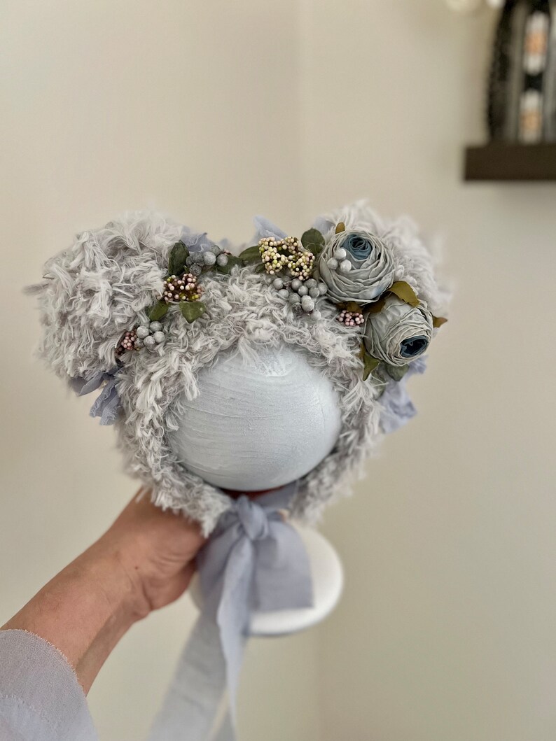 May include: A gray fuzzy baby hat with a flower crown. The crown is made of blue and white fabric flowers with green leaves and small white beads.
