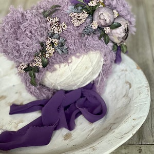 May include: A lavender fuzzy hat with a purple ribbon and a floral embellishment of white and purple flowers.