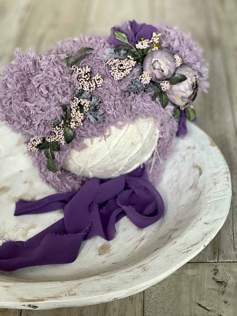 May include: A lavender fuzzy hat with a purple ribbon and a floral embellishment of white and purple flowers.