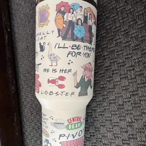 May include: A white travel mug featuring illustrations and text from the TV show "Friends." The design includes the Central Perk logo, the phrase "I'll Be There For You," and other iconic imagery from the show. The mug has a silver lid.