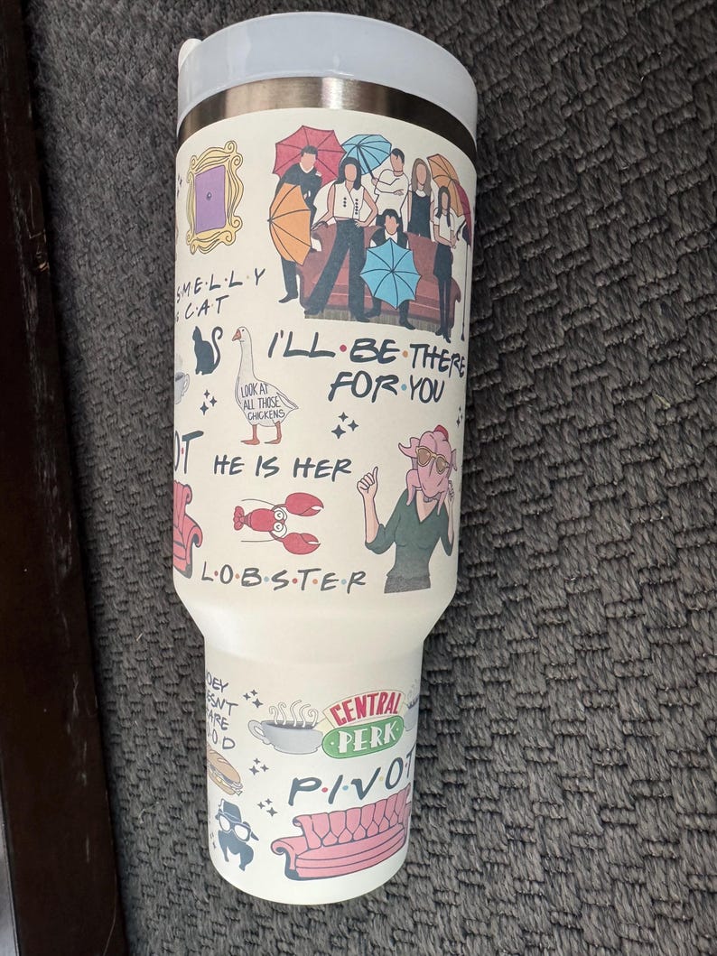 May include: A white travel mug featuring illustrations and text from the TV show "Friends." The design includes the Central Perk logo, the phrase "I'll Be There For You," and other iconic imagery from the show. The mug has a silver lid.
