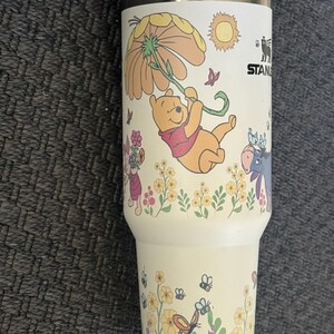 Może przedstawiać: Kremowy kubek Stanley z motywem Kubusia Puchatka. Projekt zawiera ilustracje Puchatka wiszącego na kwiatku, motyle, kwiaty i słońce. Kubek ma wnętrze ze stali nierdzewnej i białą plastikową pokrywkę.