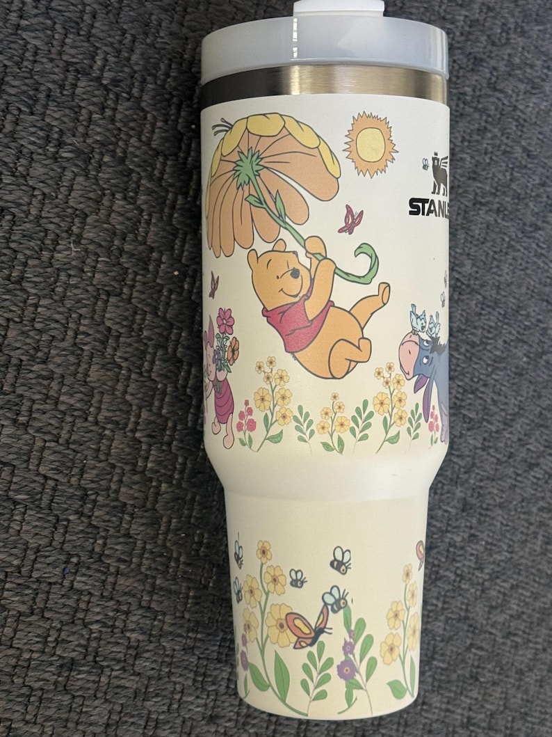 Może przedstawiać: Kremowy kubek Stanley z motywem Kubusia Puchatka. Projekt zawiera ilustracje Puchatka wiszącego na kwiatku, motyle, kwiaty i słońce. Kubek ma wnętrze ze stali nierdzewnej i białą plastikową pokrywkę.