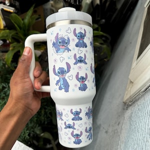 Puede incluir: Un vaso t&eacute;rmico blanco con asa, con un patr&oacute;n repetido del personaje Stitch de Lilo & Stitch. El dise&ntilde;o incluye a Stitch en varias poses, junto con flores y estrellas. El vaso tiene una tapa y un asa blancas.