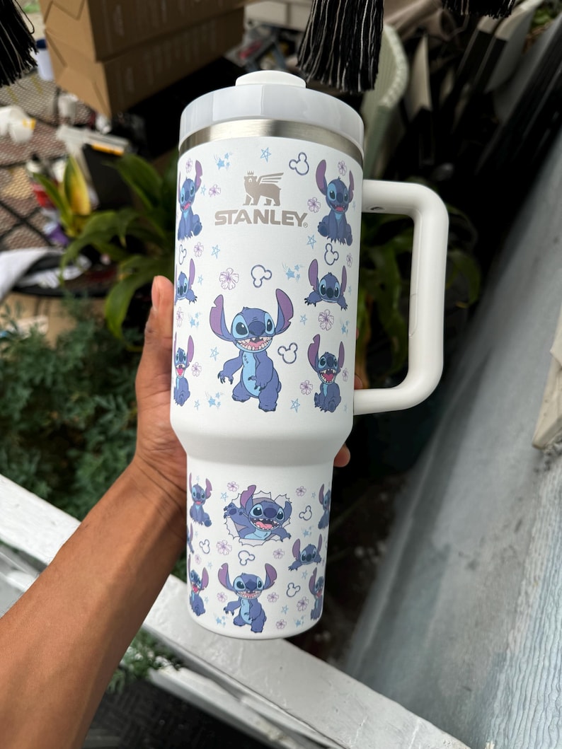 Puede incluir: Vaso Stanley blanco con dise&ntilde;o de Stitch. El vaso tiene un asa y tapa blancos, y lleva el logotipo de Stanley. El dise&ntilde;o incluye ilustraciones de Stitch y motivos florales. Adecuado para bebidas fr&iacute;as o calientes.