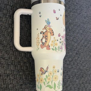 Może przedstawiać: Biały kubek podr&oacute;żny z uchwytem, z ilustracjami Tygryska, motyli, kwiat&oacute;w i innych postaci. Kubek ma szarą pokrywkę i biały uchwyt.