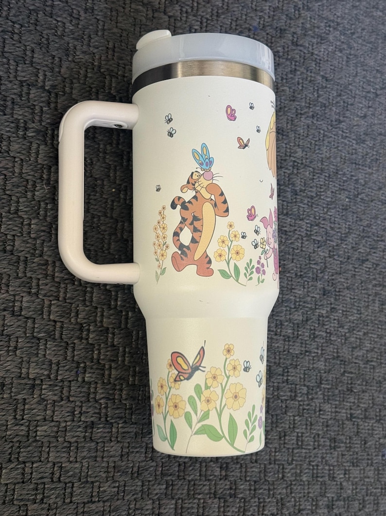 Może przedstawiać: Biały kubek podr&oacute;żny z uchwytem, z ilustracjami Tygryska, motyli, kwiat&oacute;w i innych postaci. Kubek ma szarą pokrywkę i biały uchwyt.