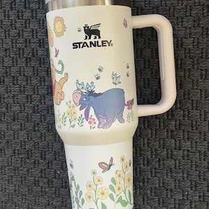 Może przedstawiać: Biały kubek podr&oacute;żny Stanley z uchwytem, z ilustracjami postaci Kubusia Puchatka, motyli i kwiat&oacute;w. Kubek ma szarą pokrywkę i biały uchwyt. Widoczne jest logo Stanley.