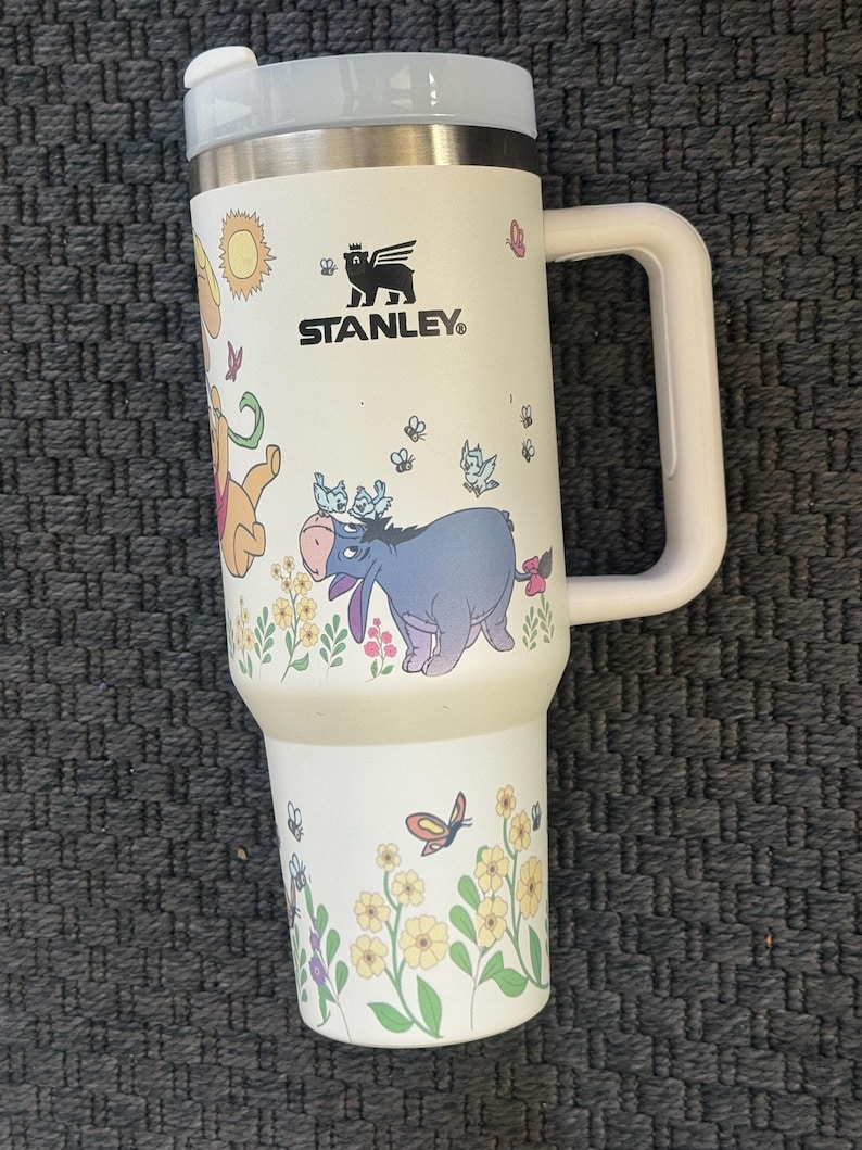 Może przedstawiać: Biały kubek podr&oacute;żny Stanley z uchwytem, z ilustracjami postaci Kubusia Puchatka, motyli i kwiat&oacute;w. Kubek ma szarą pokrywkę i biały uchwyt. Widoczne jest logo Stanley.