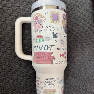 May include: White travel mug with a handle, featuring various cartoon illustrations and text from the TV show "Friends." The design includes a turkey wearing sunglasses, a purple door frame, and the words "PIVOT" and "JOEY DOESN'T SHARE FOOD."