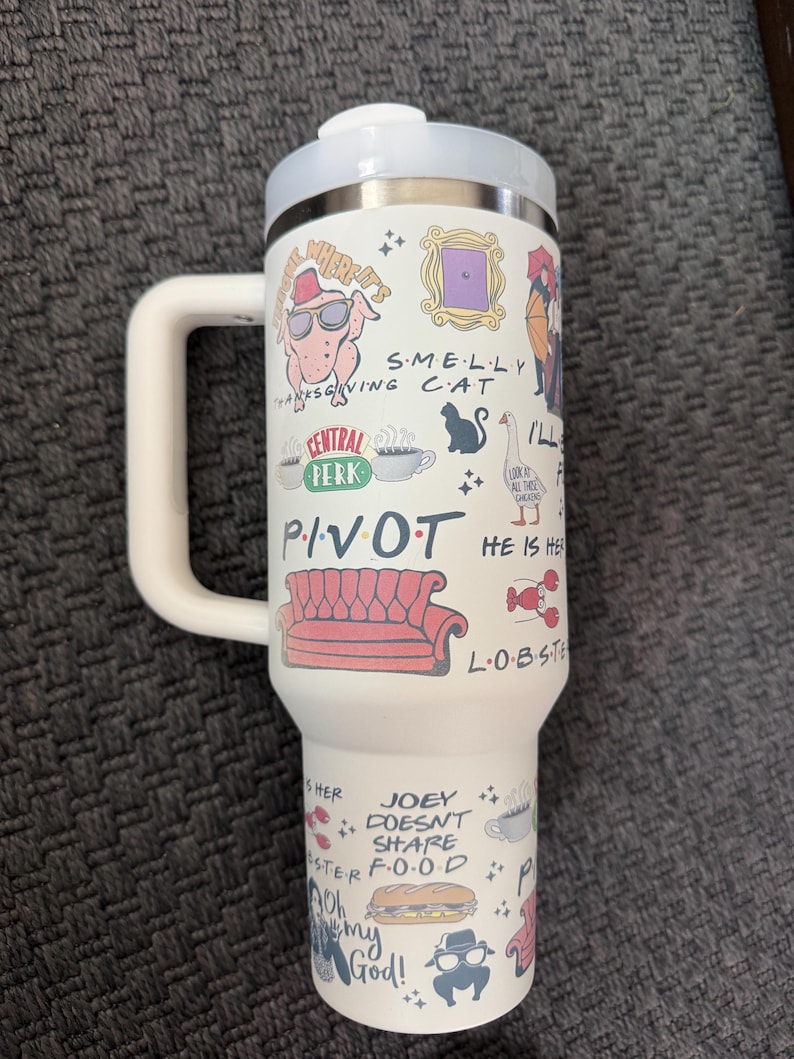 May include: White travel mug with a handle, featuring various cartoon illustrations and text from the TV show "Friends." The design includes a turkey wearing sunglasses, a purple door frame, and the words "PIVOT" and "JOEY DOESN'T SHARE FOOD."