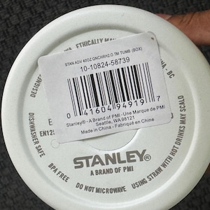 Może przedstawiać: Biała pokrywka do kubka Stanley z etykietą produktu. Etykieta zawiera kod kreskowy, informacje o produkcie oraz tekst "STAN ADV 40OZ QNCHRH2.0 TM TUMB (BOX)". Pokrywka ma r&oacute;wnież napis "DISHWASHER SAFE" i "BPA FREE".