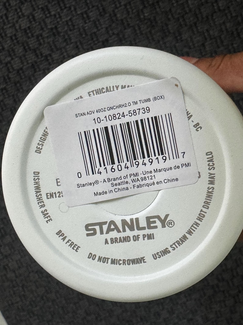 Może przedstawiać: Biała pokrywka do kubka Stanley z etykietą produktu. Etykieta zawiera kod kreskowy, informacje o produkcie oraz tekst "STAN ADV 40OZ QNCHRH2.0 TM TUMB (BOX)". Pokrywka ma r&oacute;wnież napis "DISHWASHER SAFE" i "BPA FREE".
