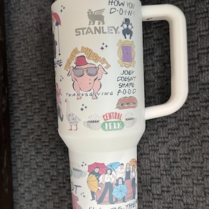 May include: White Stanley travel mug featuring various cartoon illustrations and text from the TV show Friends. The mug has a white handle and lid, with phrases like "How you doin?" and "I'll be there for you."