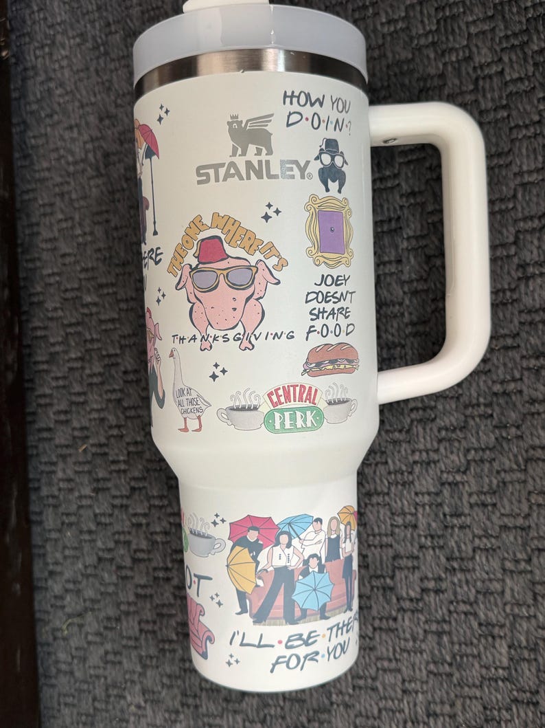 May include: White Stanley travel mug featuring various cartoon illustrations and text from the TV show Friends. The mug has a white handle and lid, with phrases like "How you doin?" and "I'll be there for you."