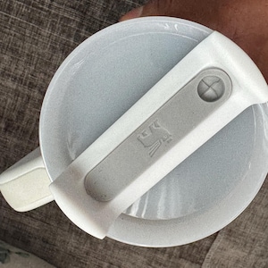 Może przedstawiać: Jasnoszara pokrywka z białym uchwytem i szarą dźwignią. Dźwignia ma mały, wytłoczony wz&oacute;r. Pokrywka wydaje się być wykonana z teksturowanego tworzywa sztucznego. Uchwyt jest przymocowany do pokrywki za pomocą zawiasu.