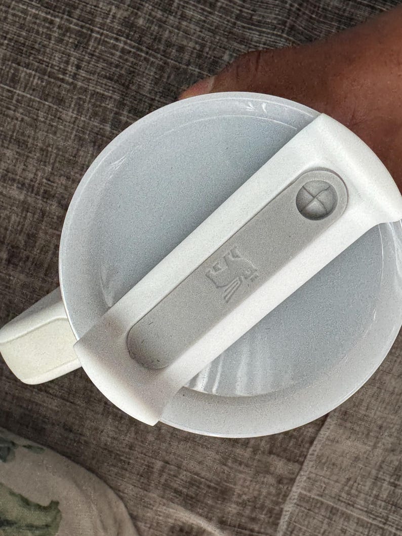 Może przedstawiać: Jasnoszara pokrywka z białym uchwytem i szarą dźwignią. Dźwignia ma mały, wytłoczony wz&oacute;r. Pokrywka wydaje się być wykonana z teksturowanego tworzywa sztucznego. Uchwyt jest przymocowany do pokrywki za pomocą zawiasu.