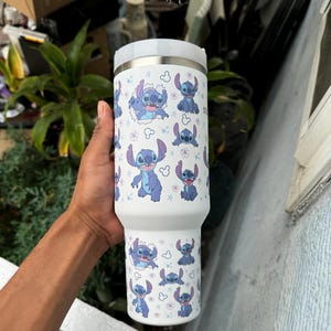 Puede incluir: Vaso t&eacute;rmico blanco con un patr&oacute;n repetido del personaje Stitch de la pel&iacute;cula Lilo & Stitch. El dise&ntilde;o incluye a Stitch en varias poses, rodeado de flores y estrellas. El vaso tiene una tapa blanca y un cuerpo de acero inoxidable.