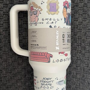 May include: A white Stanley Quencher tumbler with a handle, featuring illustrations and text from the TV show "Friends." The design includes a turkey with sunglasses, a purple door frame, and the words "SMELLY CAT."