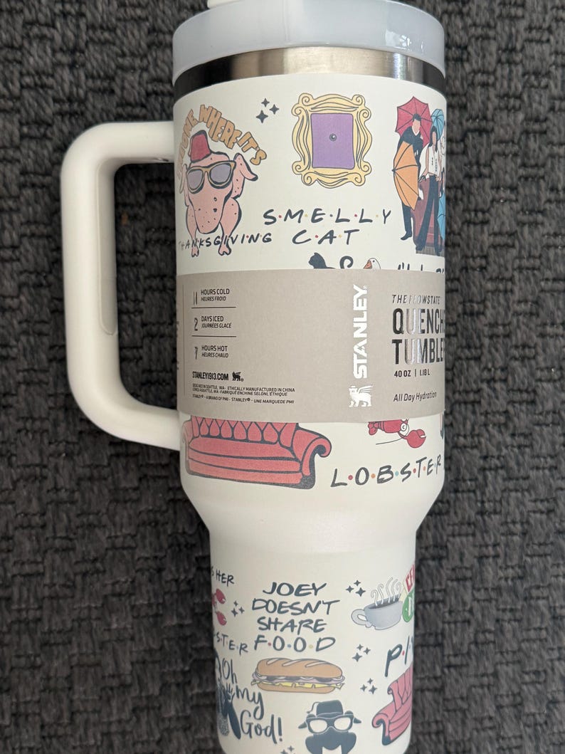 May include: A white Stanley Quencher tumbler with a handle, featuring illustrations and text from the TV show "Friends." The design includes a turkey with sunglasses, a purple door frame, and the words "SMELLY CAT."