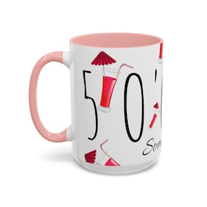 May include: White ceramic mug with a pink interior and handle. The mug features the number "50" in large black script, with illustrations of red cocktails with umbrellas. The word "Some" is written in cursive.