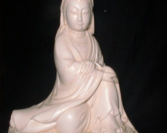 May include: A cream-colored statue of a seated figure with serene facial features, wearing draped robes and a headdress. The figure is seated on a textured base, with hands resting in the lap. The statue has a smooth, matte finish.