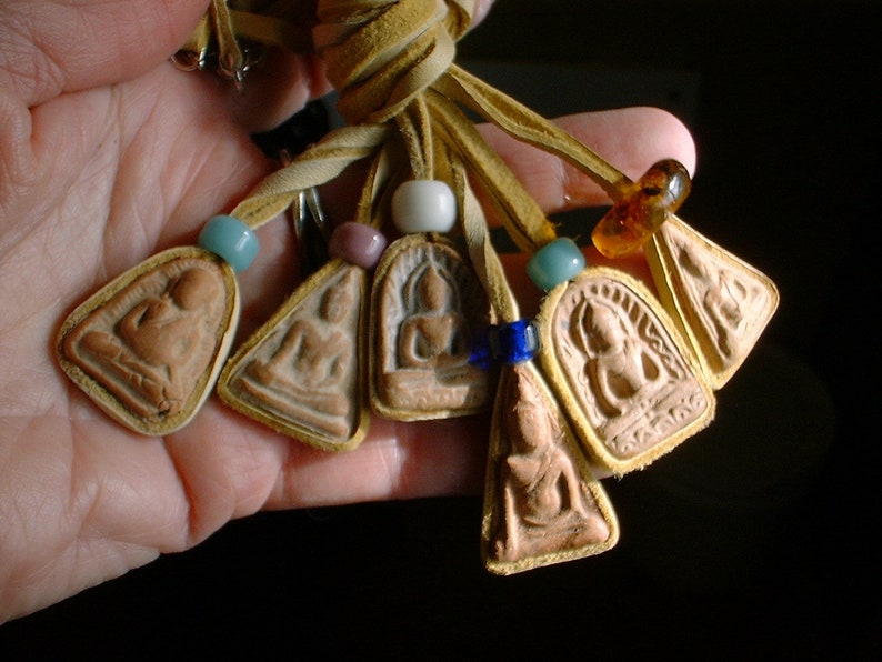 K&ouml;nnte beinhalten: Eine Halskette aus Lederband mit mehreren Anh&auml;ngern, die kleine, detaillierte Bilder von Buddha zeigen. Die Anh&auml;nger sind mit bunten Perlen verziert, darunter T&uuml;rkis, Bernstein und Wei&szlig;.