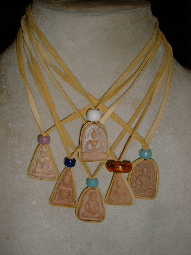 K&ouml;nnte beinhalten: Eine Halskette mit sieben Anh&auml;ngern, die kleine, braune, sitzende Buddha-Figuren zeigen. Jeder Anh&auml;nger ist an einer separaten L&auml;nge von braunem Lederband befestigt. Die B&auml;nder sind oben mit einer wei&szlig;en Perle zusammengebunden.