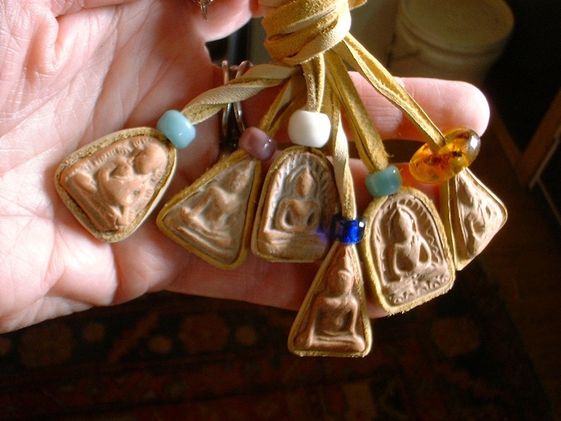 K&ouml;nnte beinhalten: Eine Lederschnur mit mehreren Anh&auml;ngern. Die Anh&auml;nger sind klein, rechteckig und aus hellem braunen Material gefertigt. Sie zeigen erhabene Bilder von Figuren. Die Schnur ist mit bunten Perlen verziert, darunter blaue, wei&szlig;e und bernsteinfarbene.