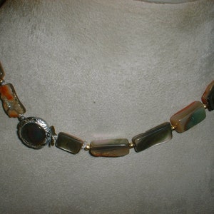 May include: A necklace featuring rectangular, multi-colored stones in shades of green, brown, and orange. The stones are strung together with small gold-colored beads. A silver-toned, oval-shaped clasp with a dark stone is visible.
